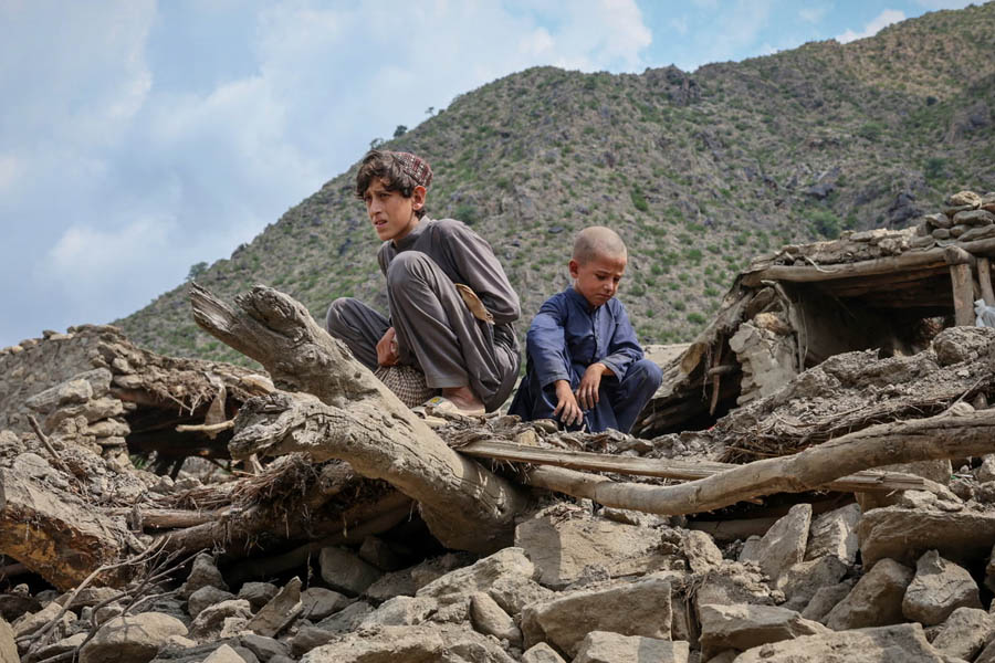 Two young Afghan boys sitting on rubble of a collapsed house in Kunar province after the deadly earthquake, symbolizing destruction and loss.