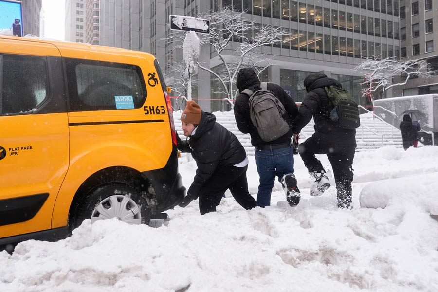 A man helping push a New York City yellow taxi stuck in deep snow during a Monday snowstorm, with snow-covered streets and low visibility in the background.