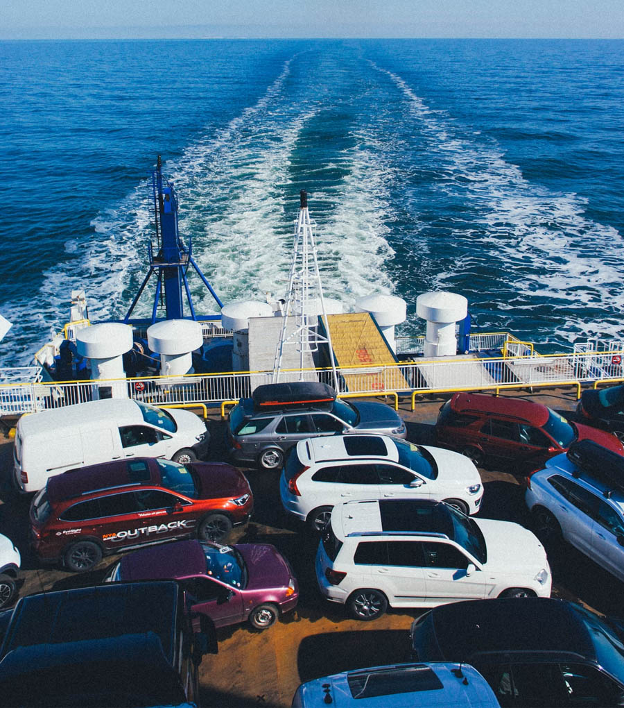 Hundreds of new vehicles packed tightly on a massive cargo ship at port, ready for transport to European markets, with shipping containers and cranes visible in the background.